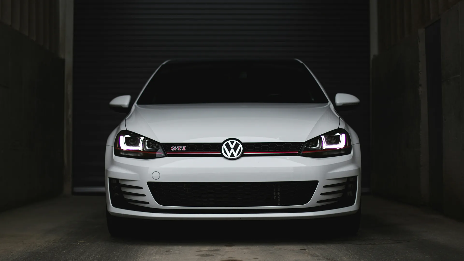 White Volkswagen Golf GTI Mk7 head-on in a dark garage with headlights lit and red grille stripe visible
