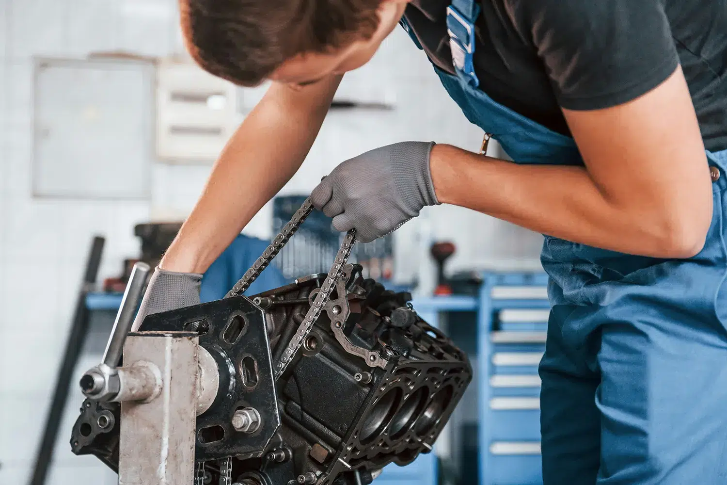 Mechanic in blue overalls fitting a timing chain to a bare engine block on a workshop stand
