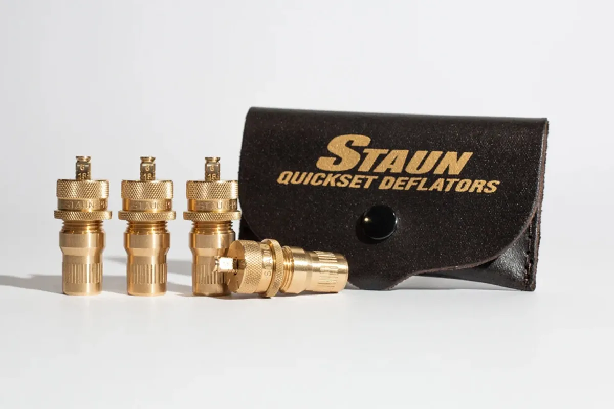 Four brass Staun Quickset tyre deflators standing beside their branded leather storage pouch on a white background