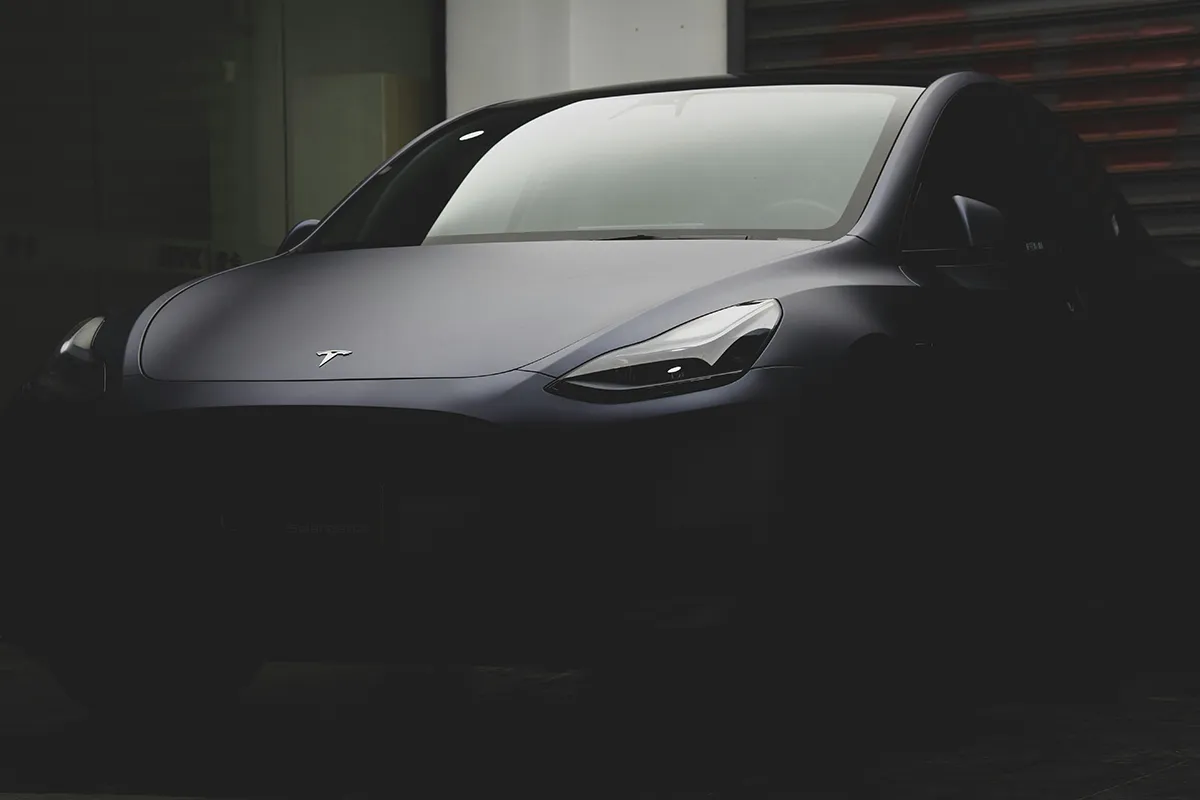 Dark grey Tesla emerging from shadow in a dimly lit garage, front three-quarter view