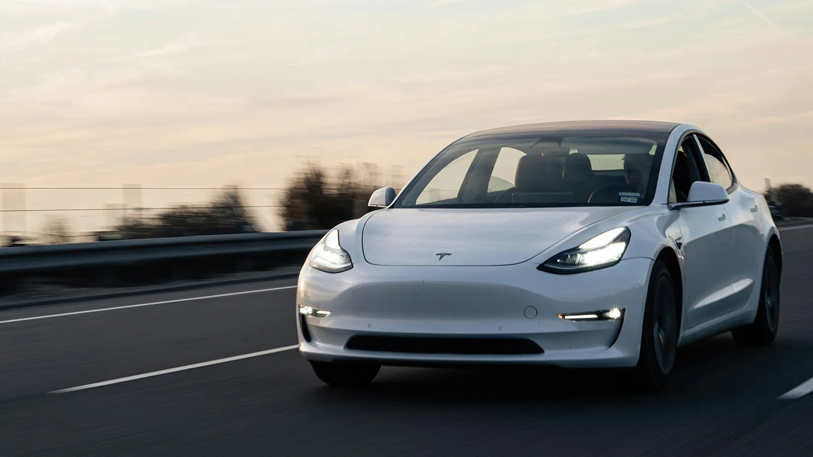 White Tesla Model 3 driving on a highway at dusk, front three-quarter view in motion
