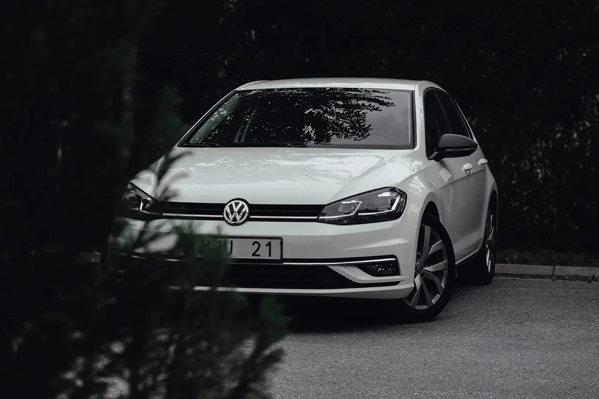 White Volkswagen Golf Mk7.5 with LED headlights lit, parked against dark forest, front three-quarter view