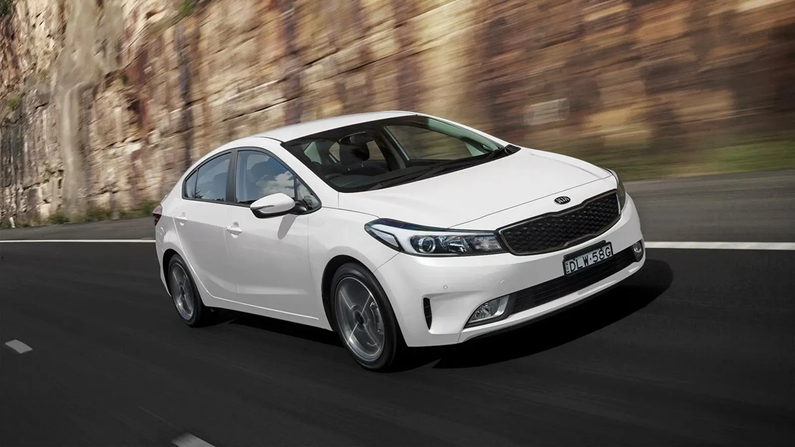 White Kia Cerato YD sedan driving past a sandstone rock cutting on an Australian highway