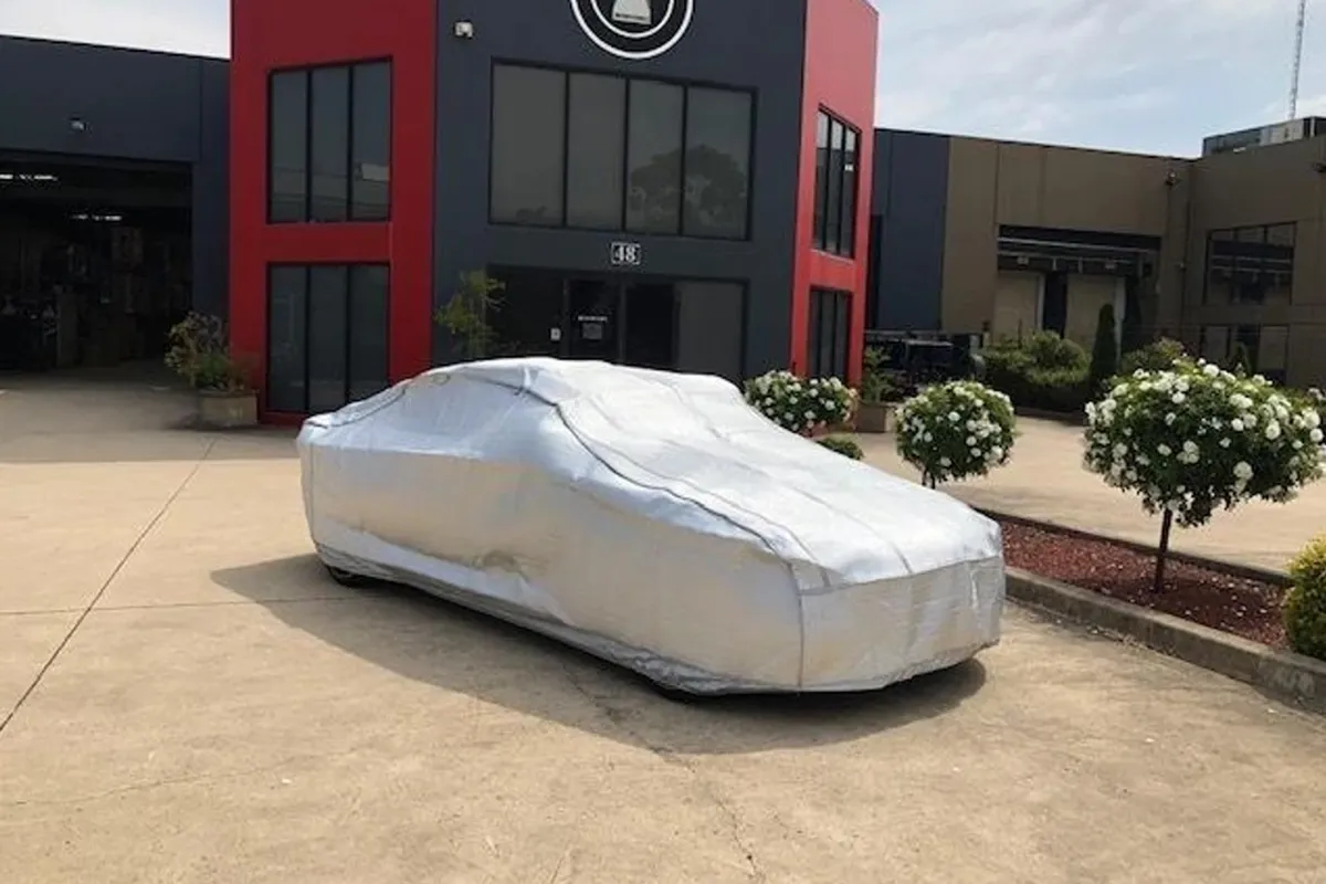 Silver weatherproof car cover fitted over a low sedan parked on concrete outside a red industrial building