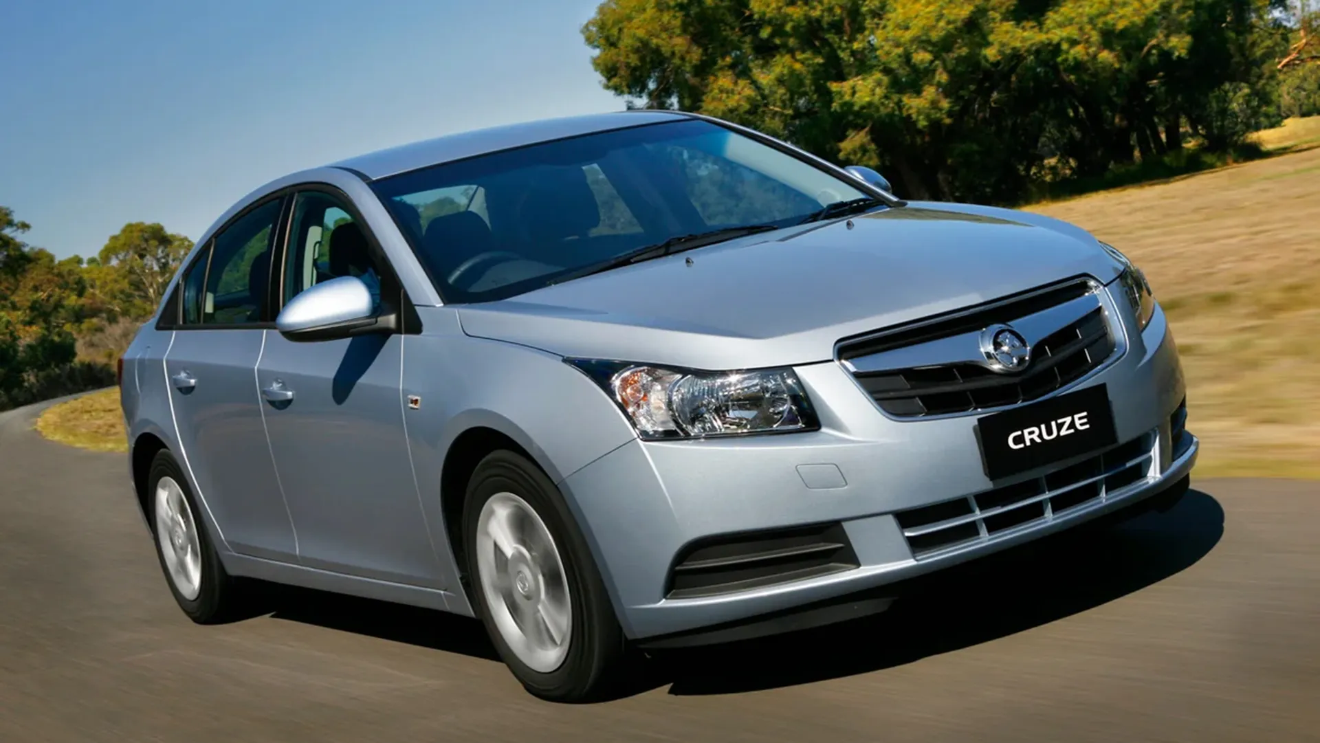 Silver first-generation Holden Cruze sedan driving on a country road lined with gum trees