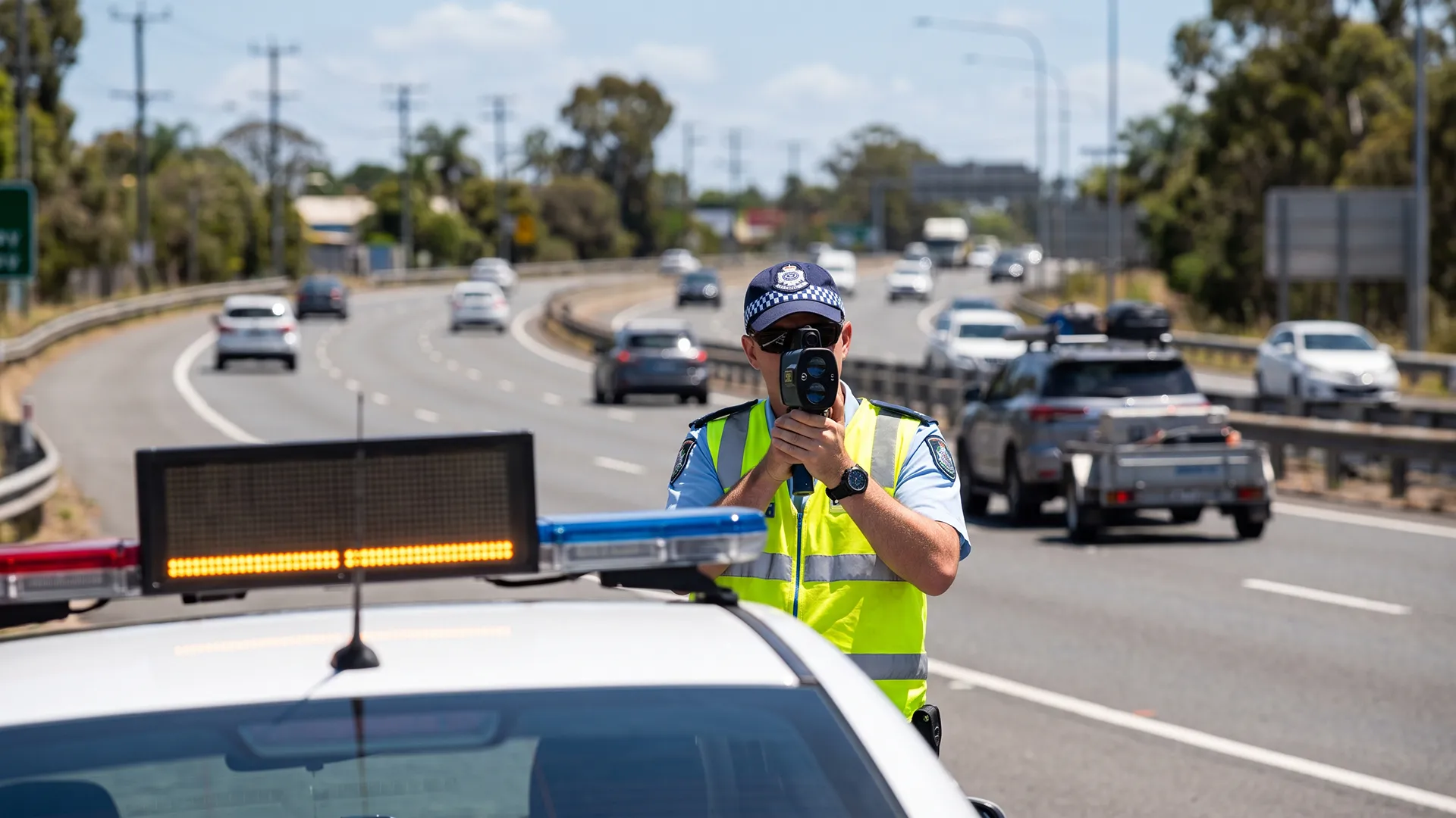 australia-police-radar-gun-traffic Highway patrol officer in a hi-vis vest aiming a laser speed gun at motorway traffic from behind a patrol car