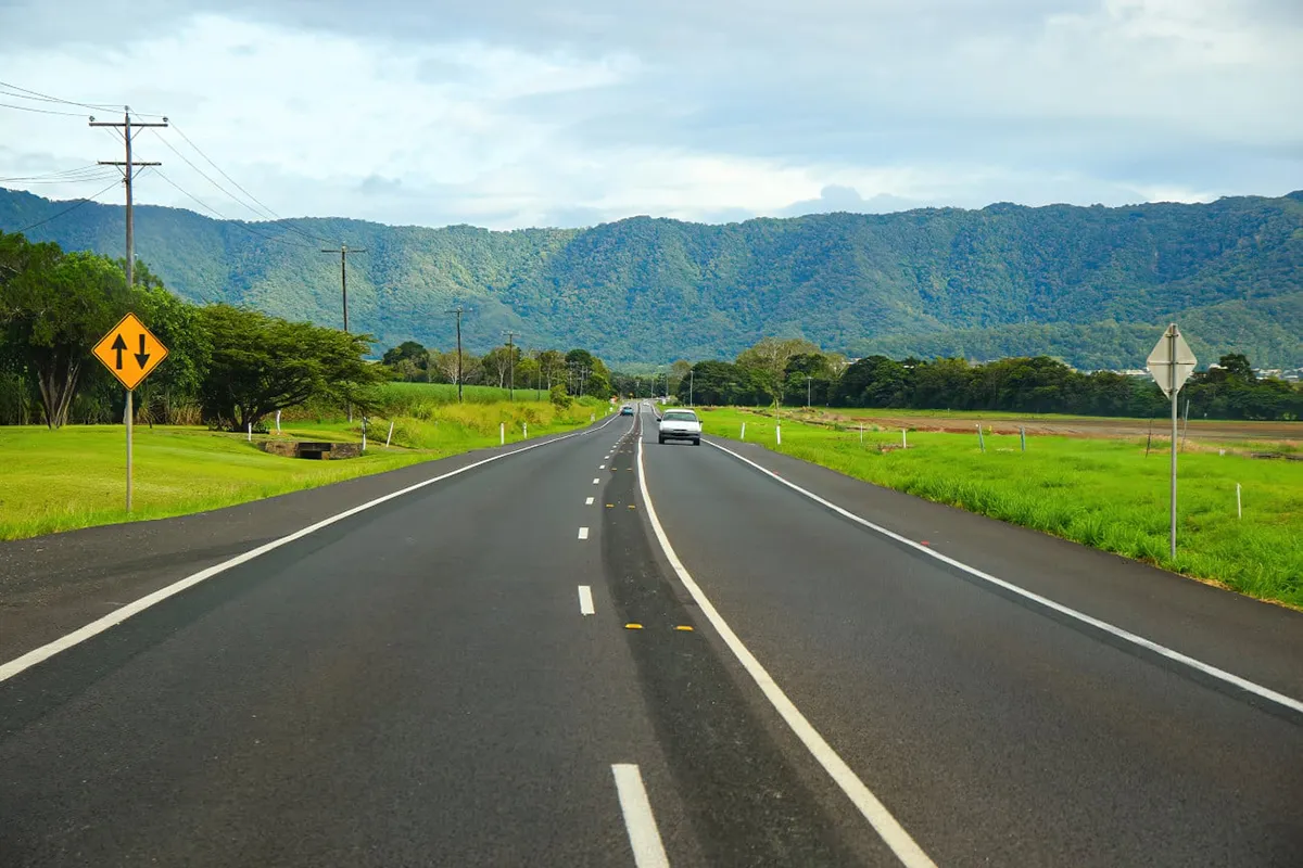 Two-lane highway through sugar cane country with rainforest-covered ranges behind in tropical north Queensland