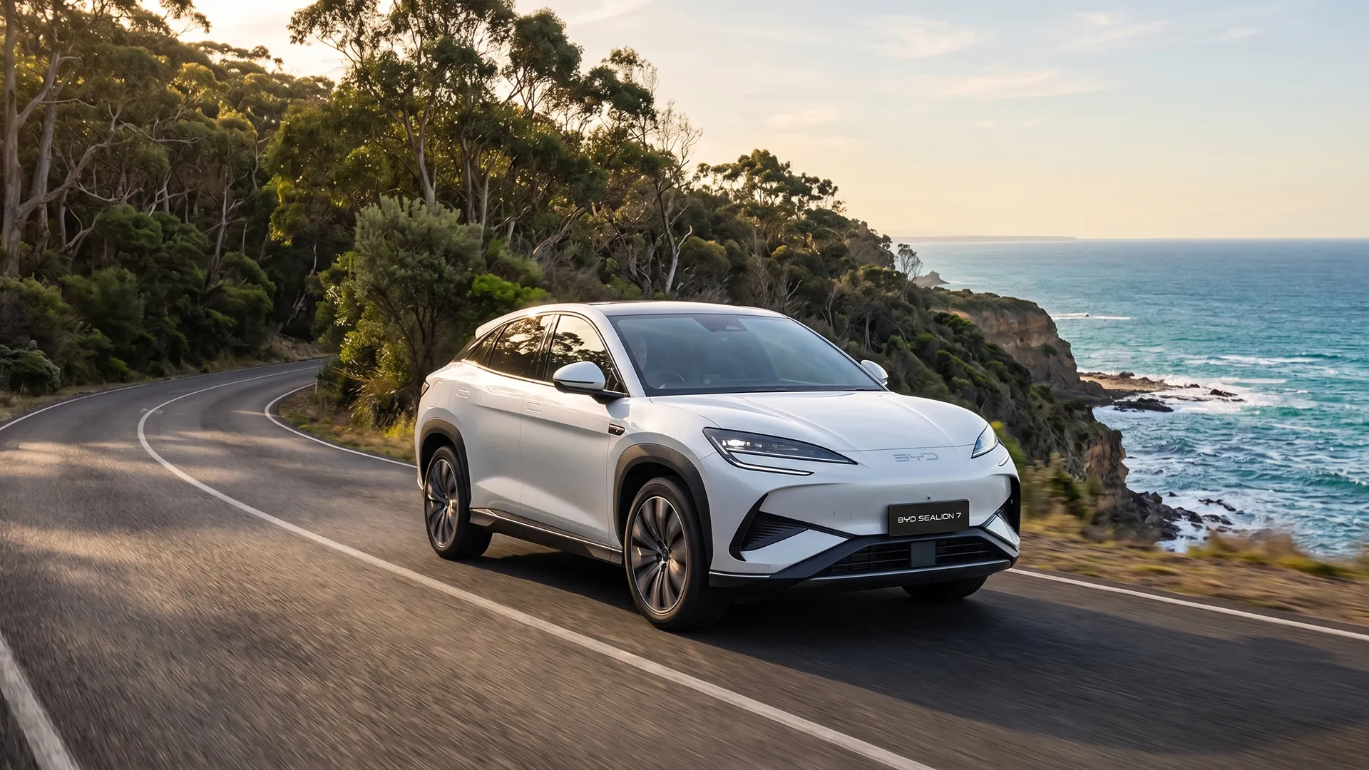 byd-sealion-7-driving-great-ocean-road-trip White BYD Sealion 7 electric SUV on a winding coastal road above ocean cliffs at sunset
