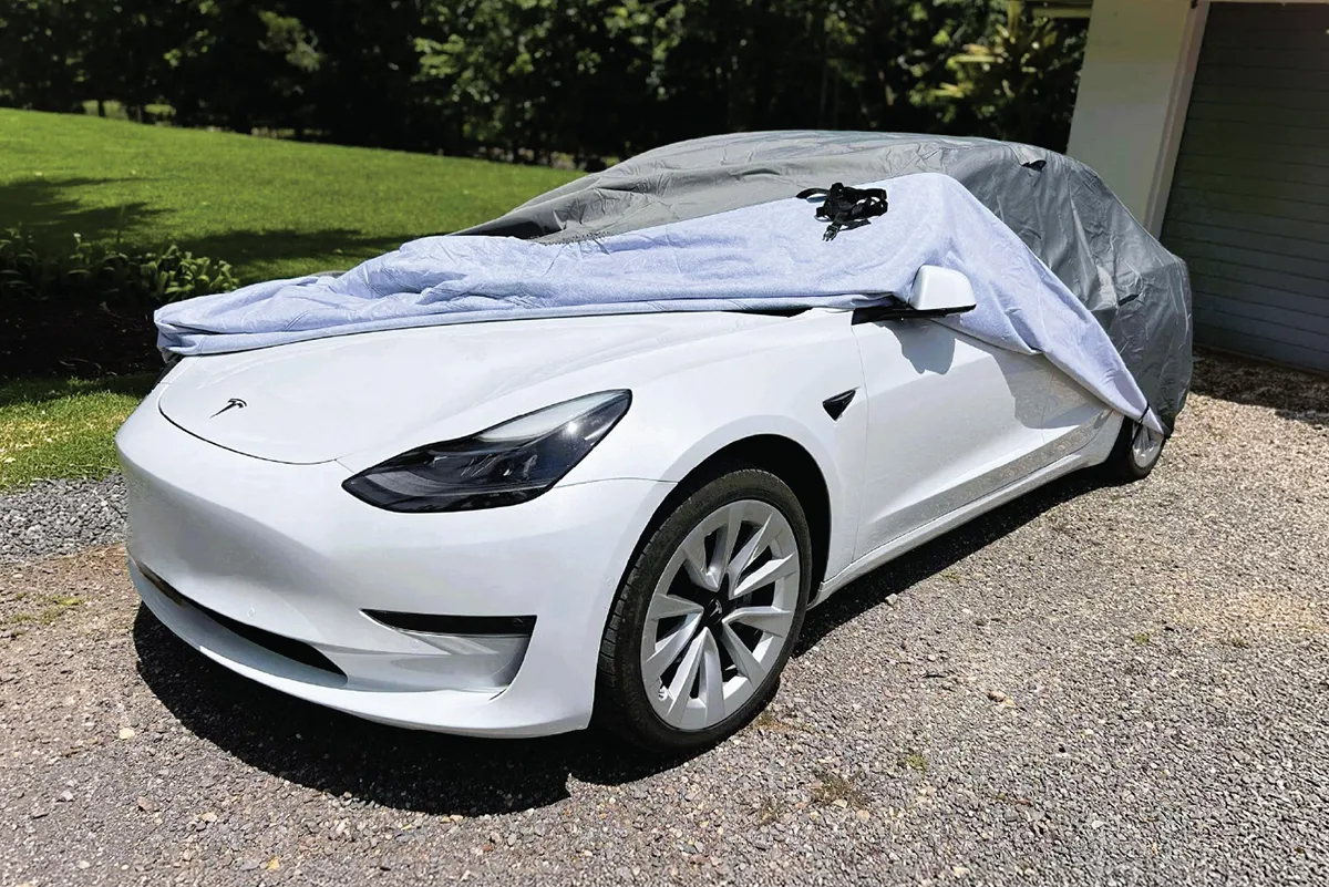 Grey car cover pulled halfway back off a white Tesla Model 3 on a gravel driveway