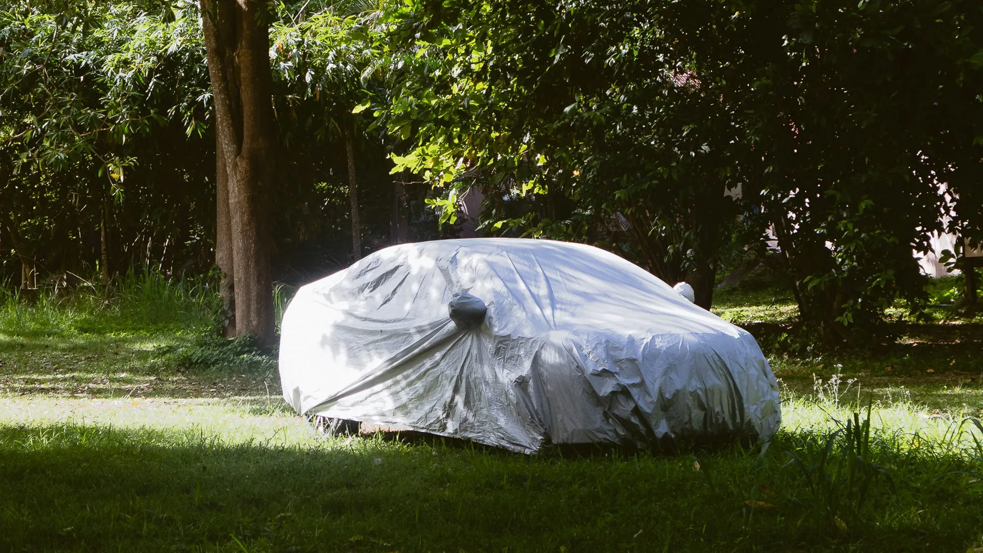 car-outdoor-car-cover Loose-fitting outdoor car cover sagging over a hatchback parked on grass beneath shady trees