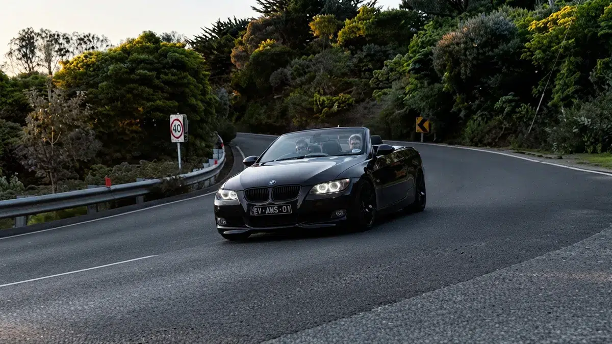 Front-three-quarter view of a black BMW E93 convertible driving through a sweeping bend on the Esplanade, Mornington Peninsula, with illuminated angel-eye headlights and two occupants inside.
