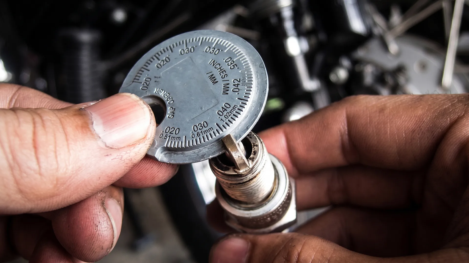 A coin-style gap gauge tool pressed against a spark plug electrode, the type often used incorrectly on fine-wire tips