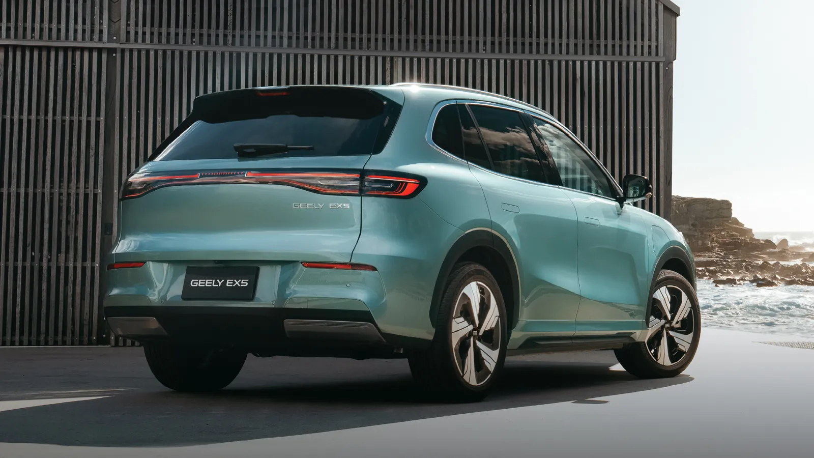 Teal Geely EX5 parked beside a corrugated shed near the coast, rear three-quarter view