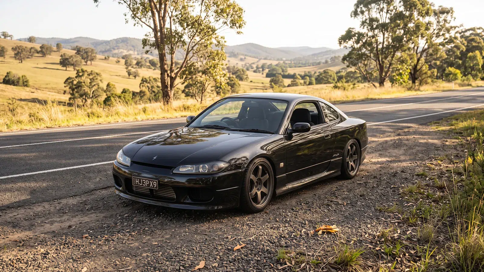 Black Nissan Silvia S15 parked on a gravel roadside with rolling farmland and hills behind