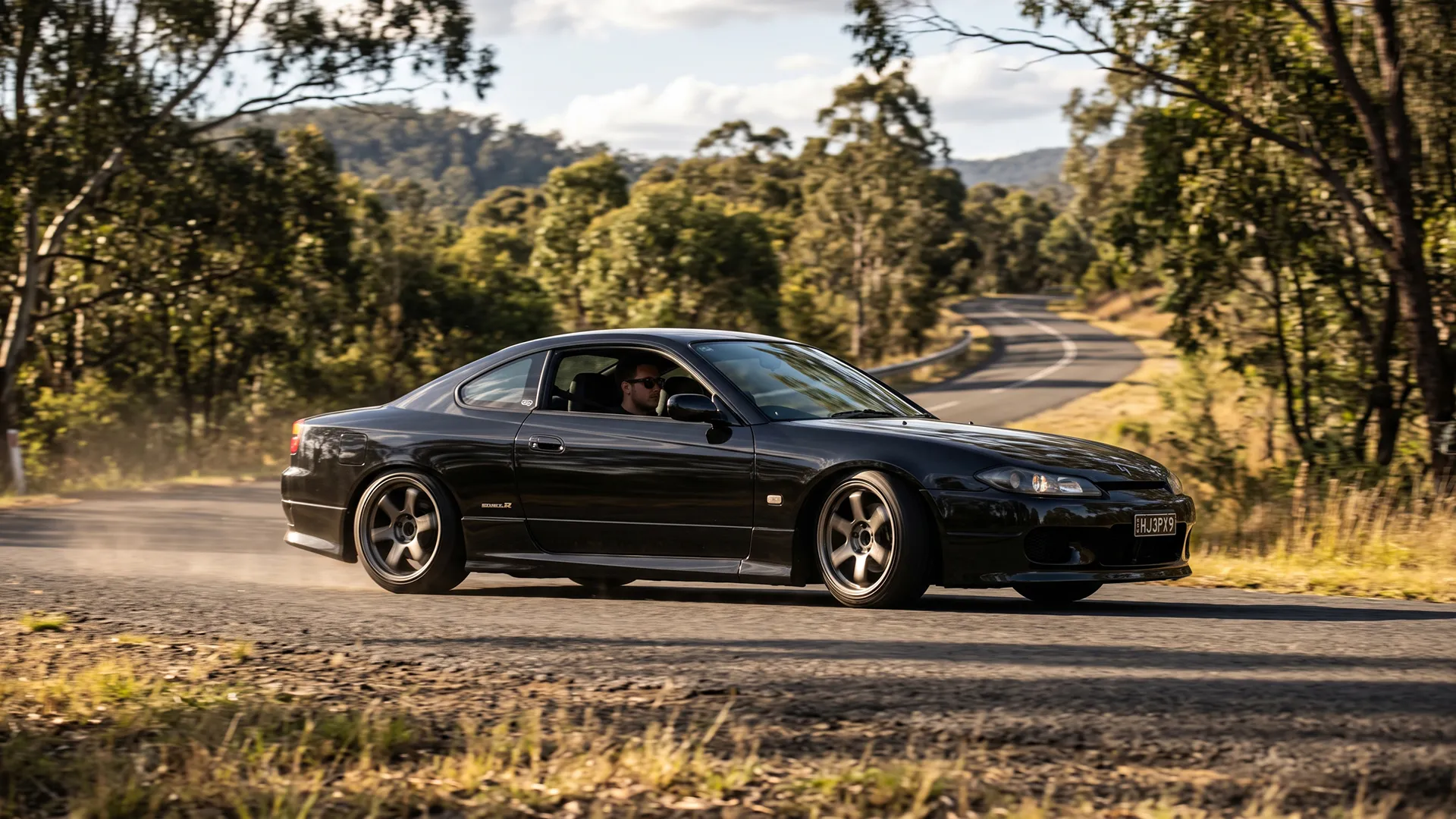 Black Nissan Silvia S15 on a curving forest road with the driver visible through the side window
