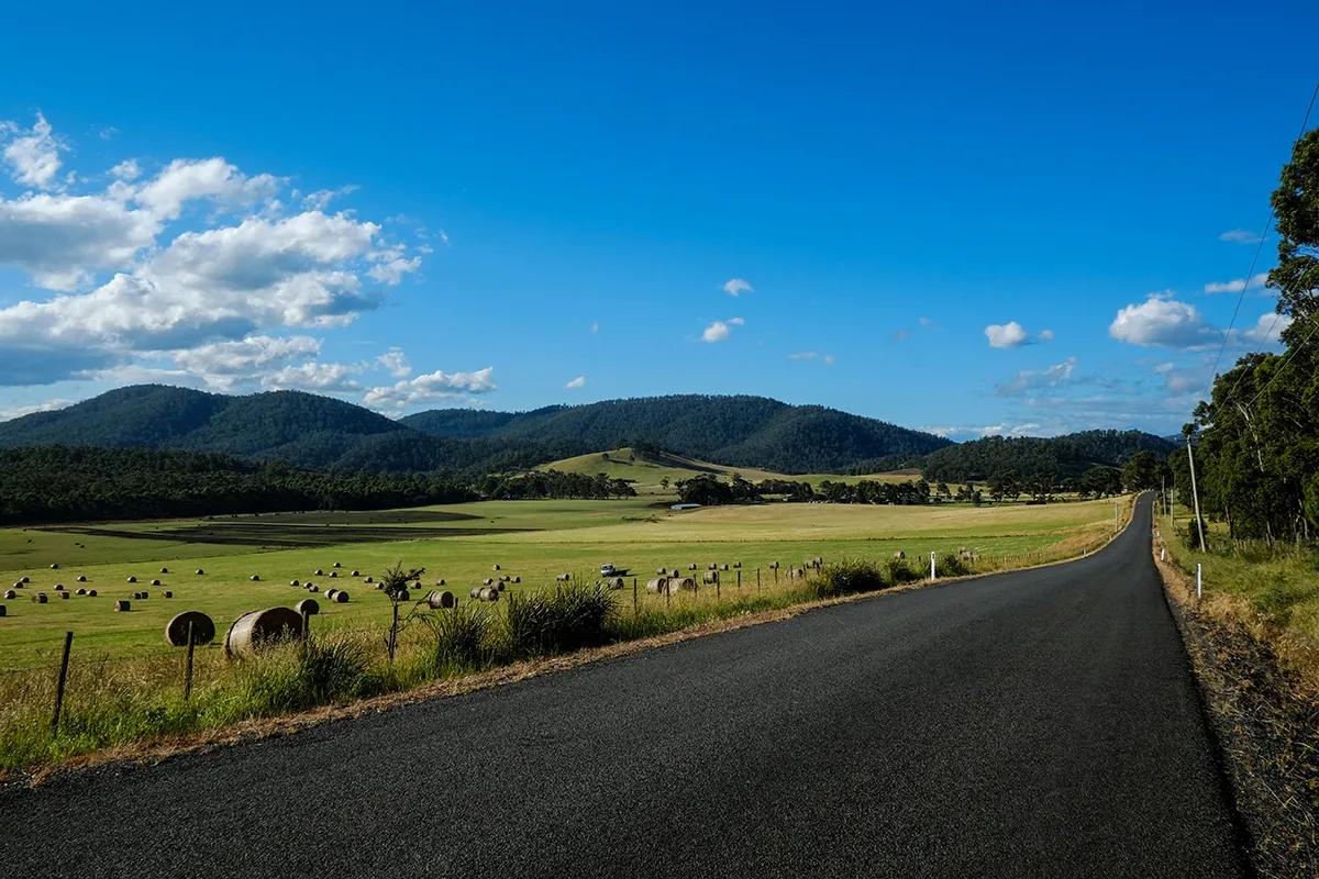 Quiet sealed country road beside hay bales and green paddocks with forested hills in rural Tasmania