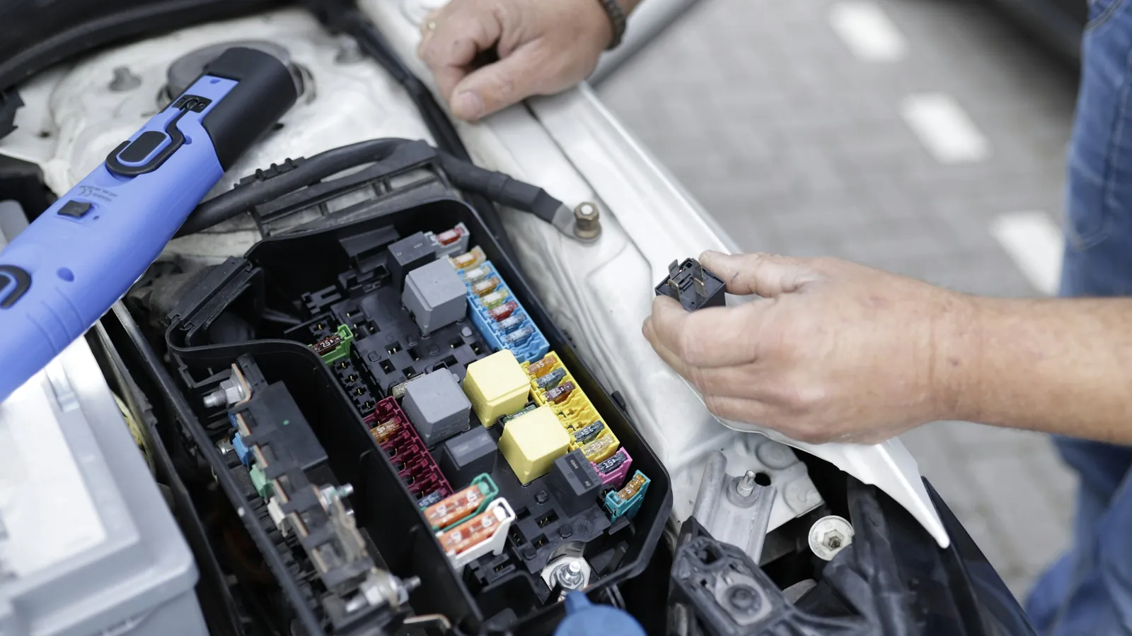 Technician working on a car's fuse box and electrical system with a diagnostic tool beside the battery