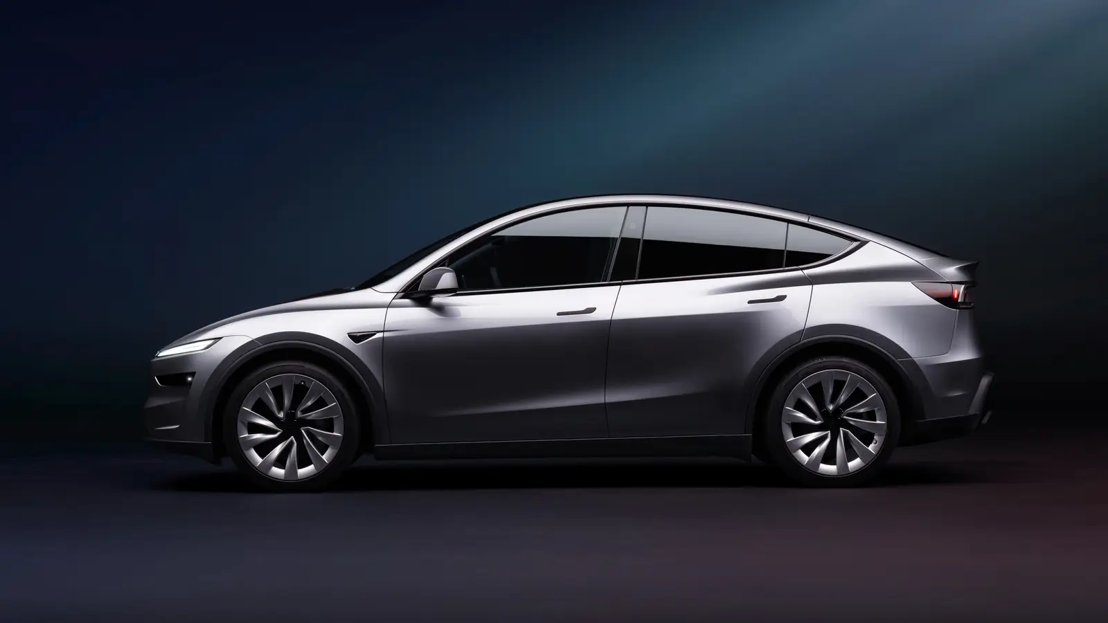 Side profile of a grey 2026 Tesla Model Y Premium in a dark studio showing its roofline and wheelbase