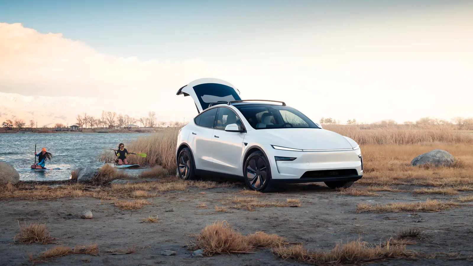 White Tesla Model Y parked by a lake with its powered tailgate open and paddleboarders in the water