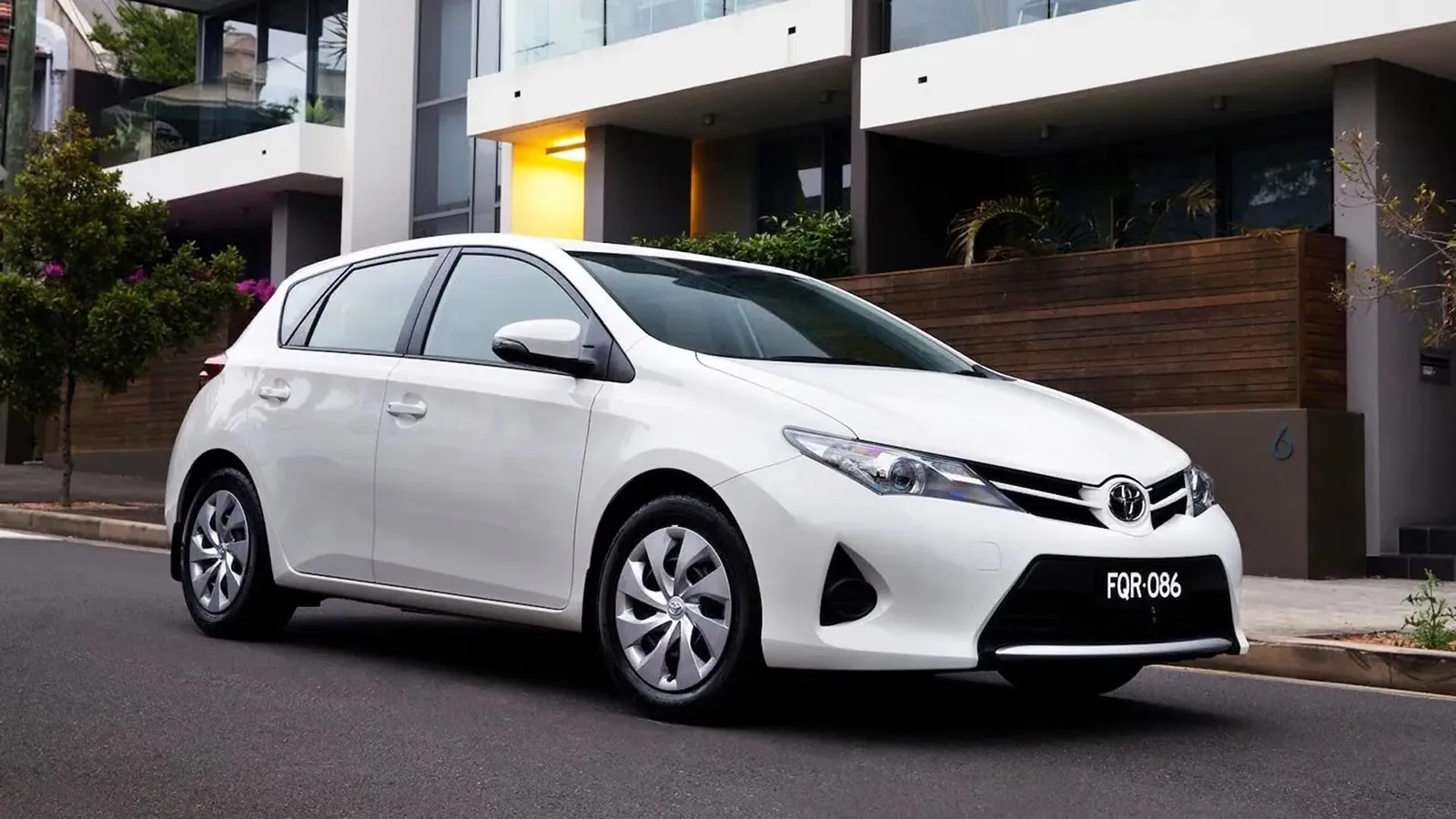White Toyota Corolla Ascent hatchback with steel wheel covers parked on a street outside modern apartments