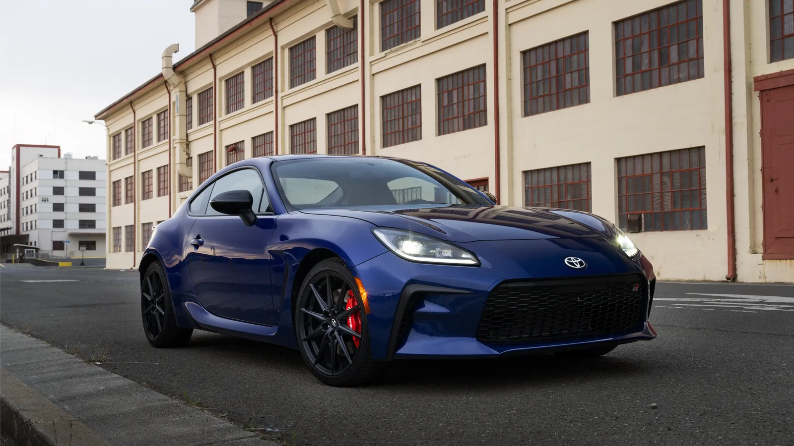 Blue Toyota GR86 sports coupe parked in front of an old factory building, front three-quarter view