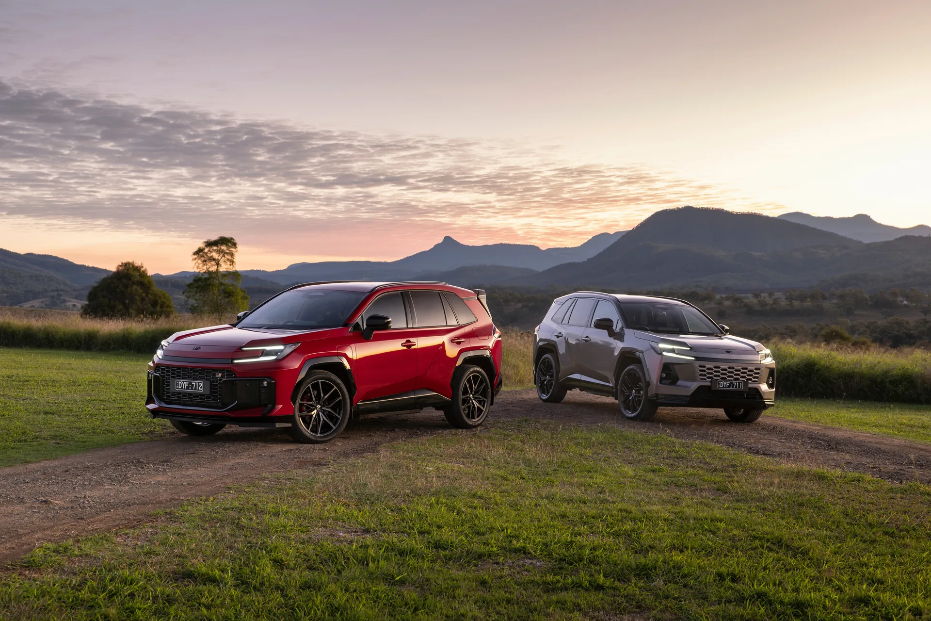 Two sixth-generation 2026 Toyota RAV4 SUVs parked on grass with mountains behind at sunset