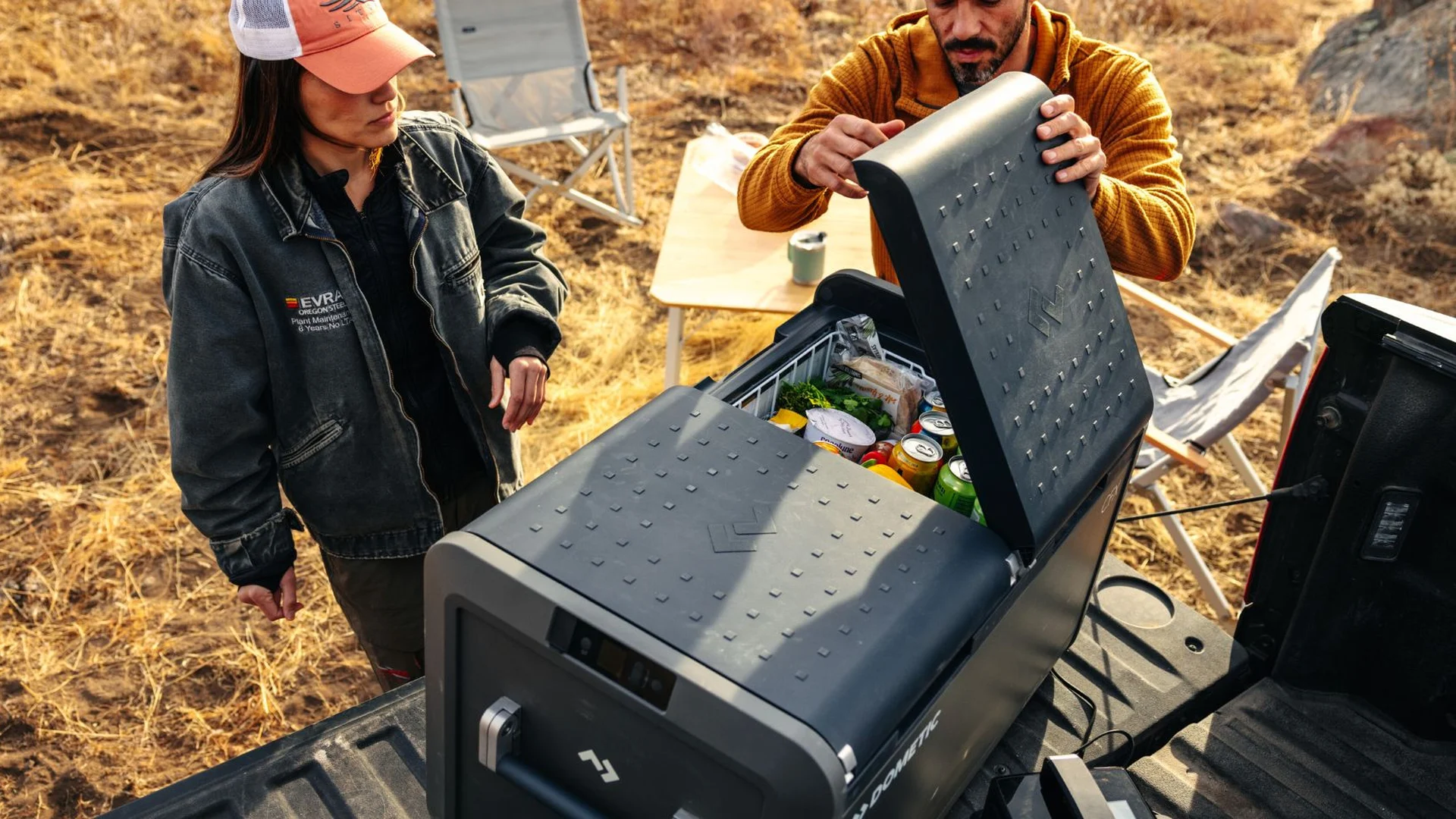 Dometic CFX5 dual-zone fridge on a ute tray Two campers loading a Dometic CFX5 dual-zone 12V fridge freezer on the tray of a ute at a dry grassy campsite