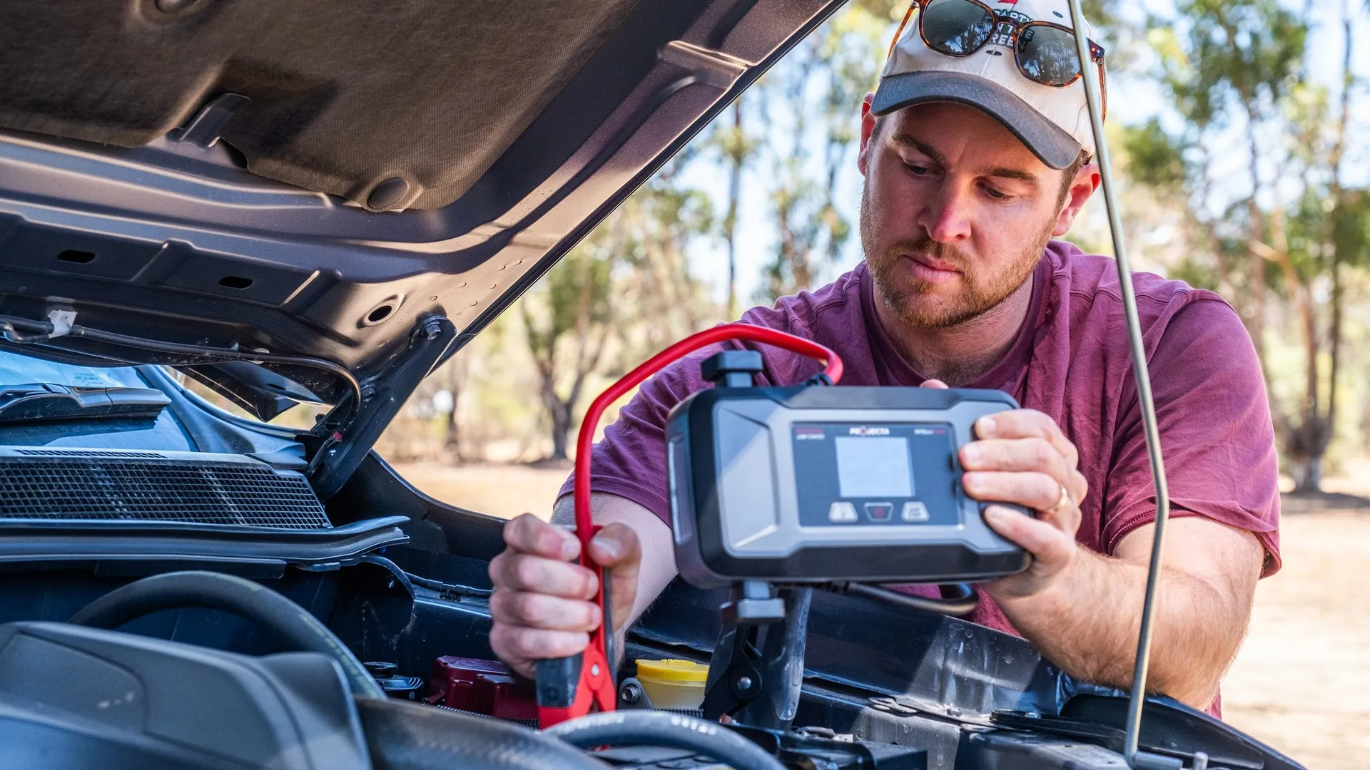 Projecta Intelli-Start IS1400 jump starter in use on a 4WD, best jump starter Australia Man connecting a Projecta Intelli-Start IS1400 jump starter to a 4WD battery under an open bonnet in the bush