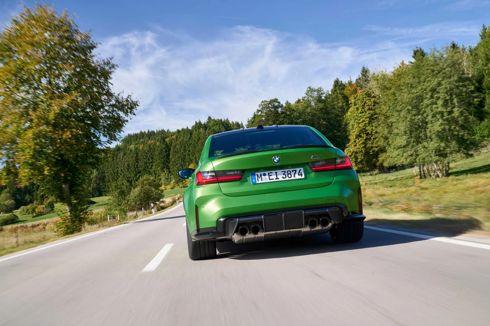 Green 2025 BMW M3 Competition sedan driving away on a country road, showing its quad exhausts and diffuser
