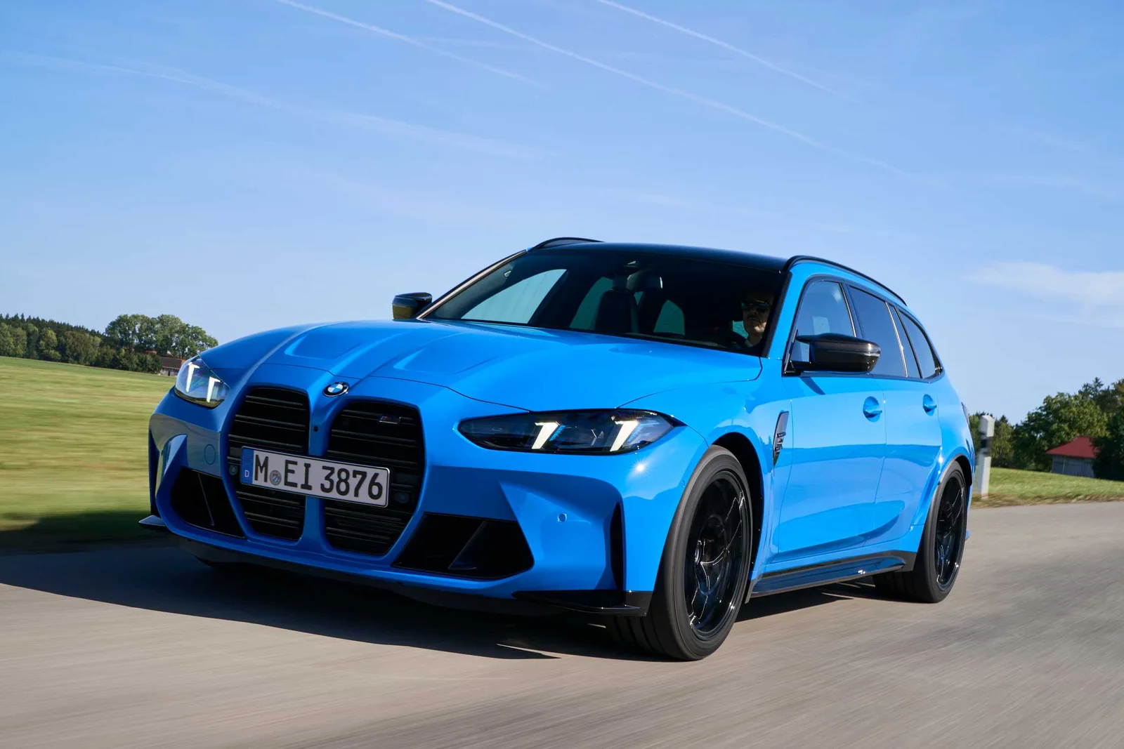 Blue 2025 BMW M3 Competition Touring wagon driving on a rural road, front three-quarter view