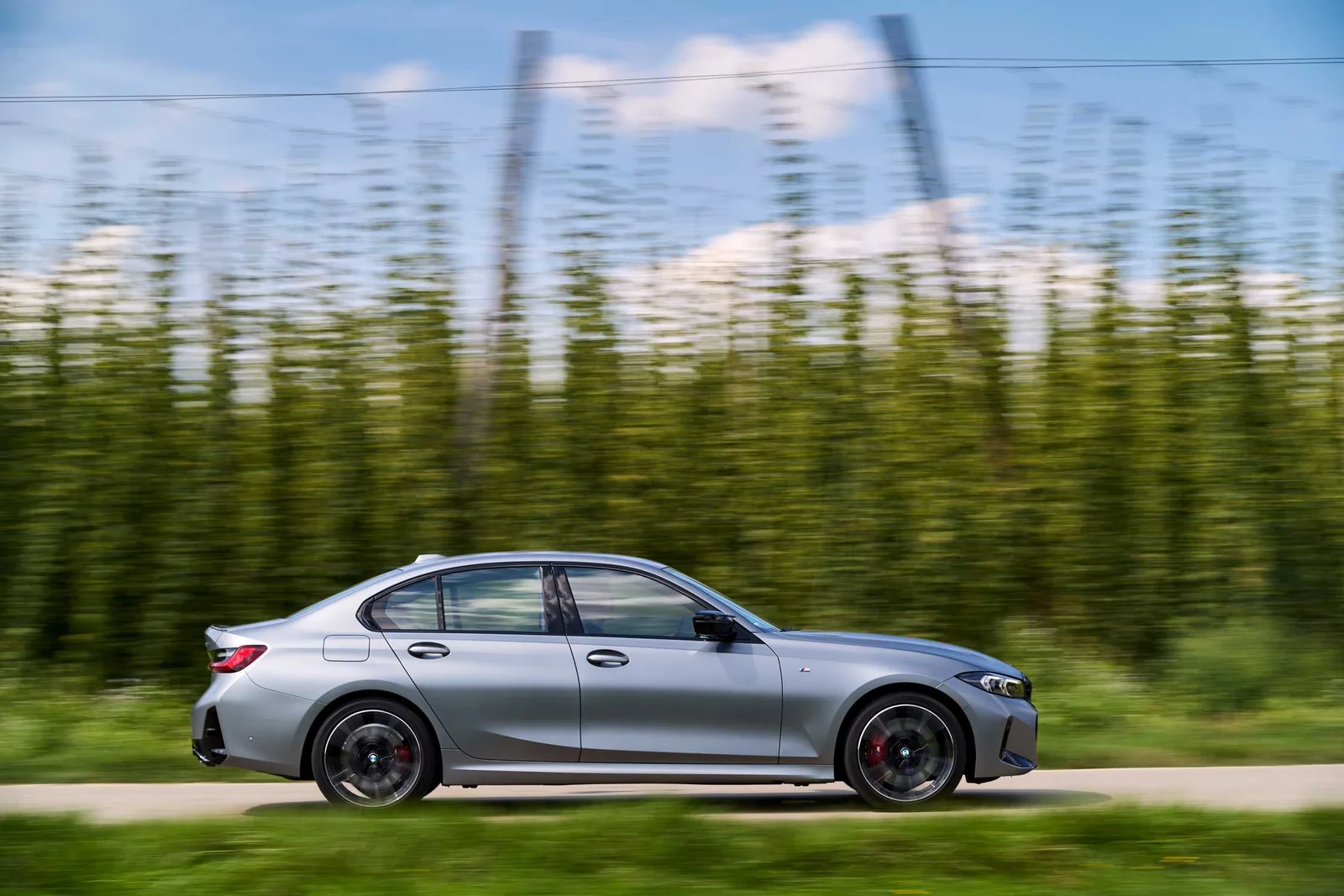 Grey BMW M340i xDrive sedan driving past a hedge, side profile with motion blur on its 19-inch wheels