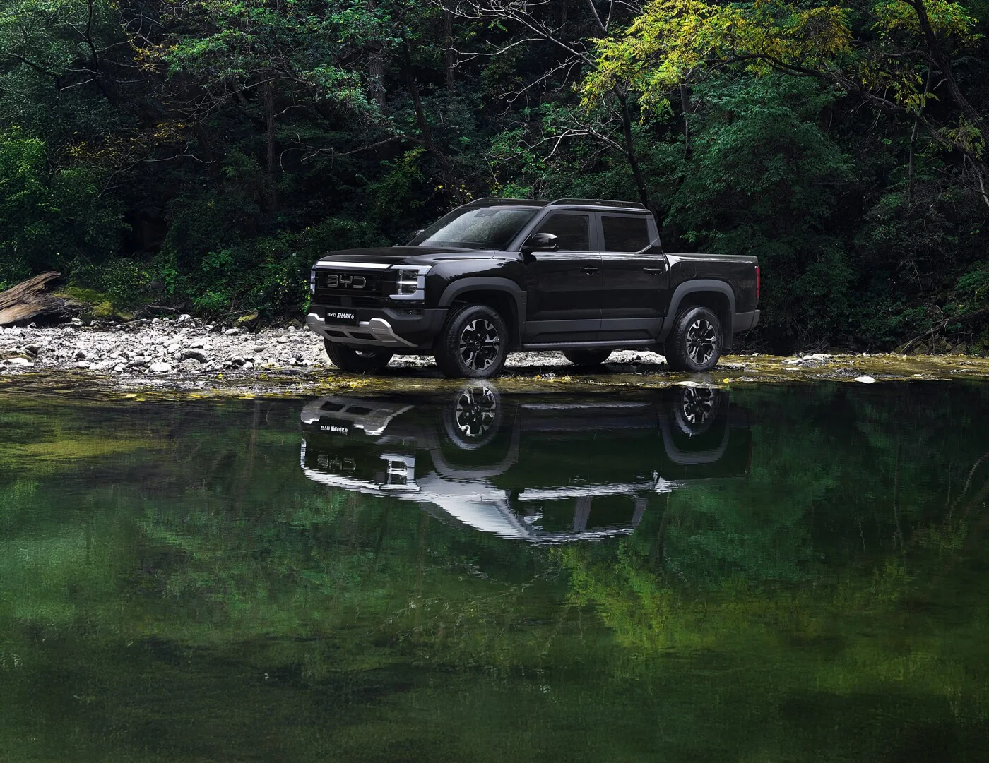 Black BYD Shark 6 parked on a rocky creek bank in forest with its reflection in the water