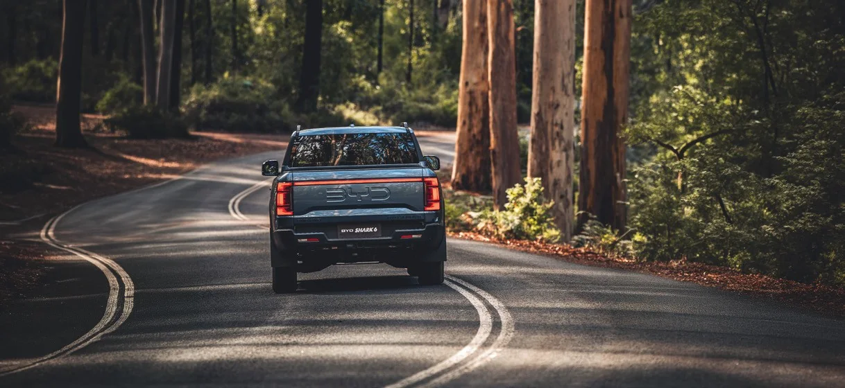 Rear view of a BYD Shark 6 on a winding road through tall eucalypt forest, showing the tailgate light bar