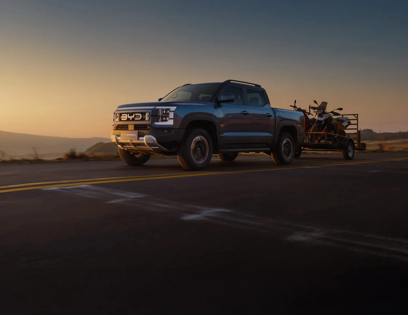 BYD Shark 6 towing a trailer carrying two motorbikes along a bitumen road at sunset