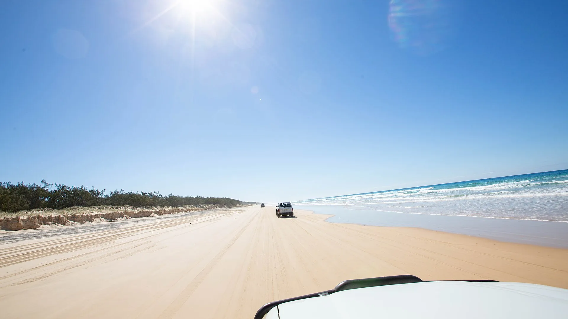 4WD driving 75 Mile Beach, K'gari View over the bonnet of a 4WD driving the hard sand of 75 Mile Beach on K'gari with another 4WD ahead and surf to the right