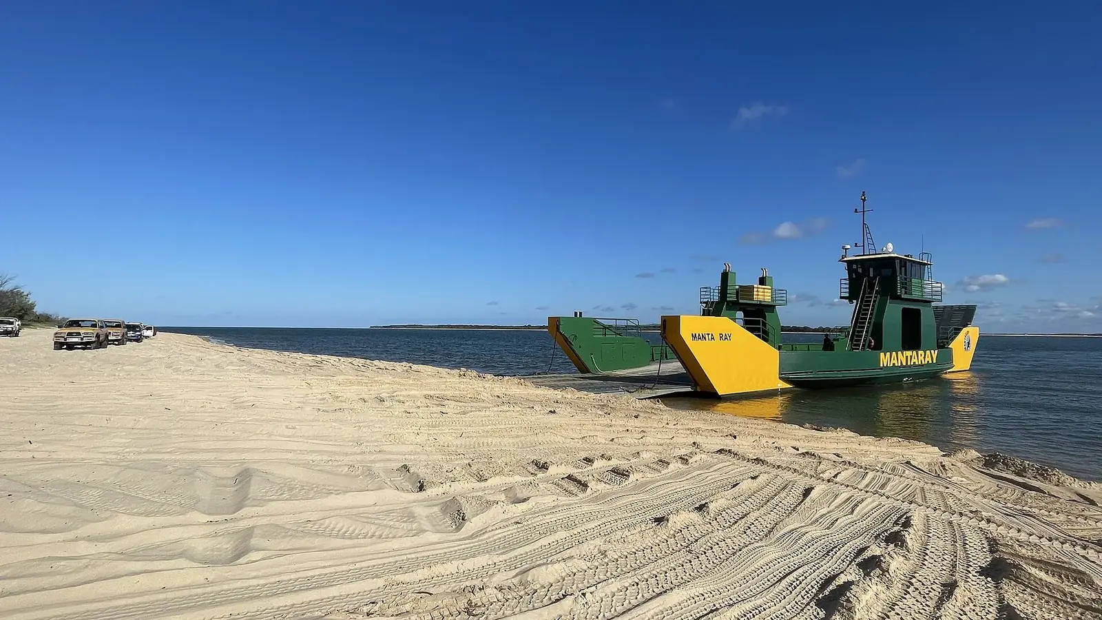 Green and yellow Mantaray vehicle barge nosed onto the sand at the Hook Point landing on K'gari, tyre tracks in the foreground