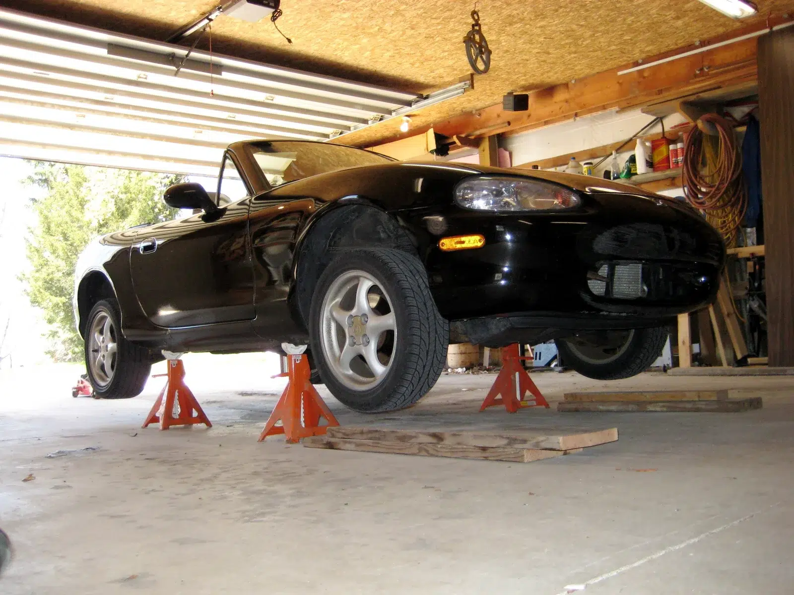 Black Mazda MX-5 roadster raised on four jack stands in a home garage with a workbench behind