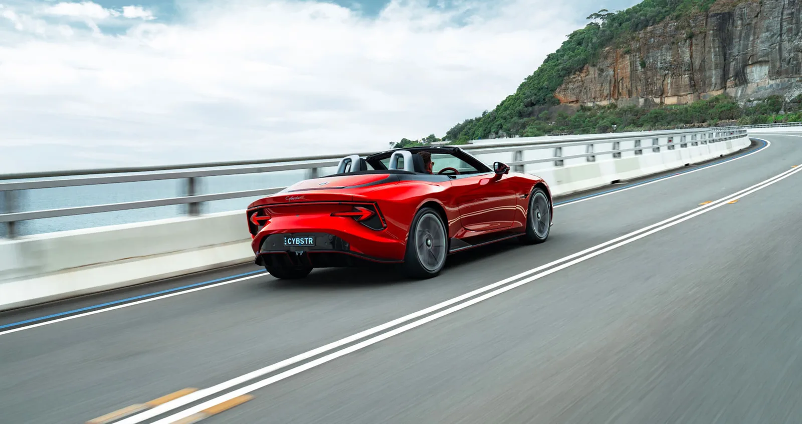 Rear three-quarter view of a red MG Cyberster driving on a coastal road with the roof down