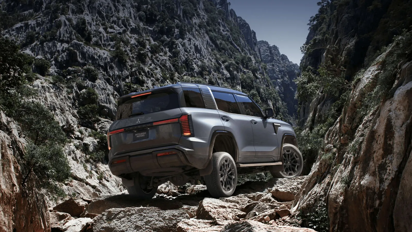 Grey 2027 Mitsubishi Pajero climbing a rocky gorge track between cliffs, rear three-quarter view