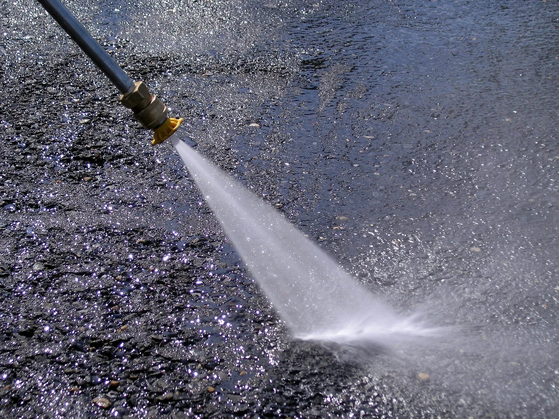 Brass pressure washer nozzle firing a narrow, concentrated jet of water onto wet asphalt