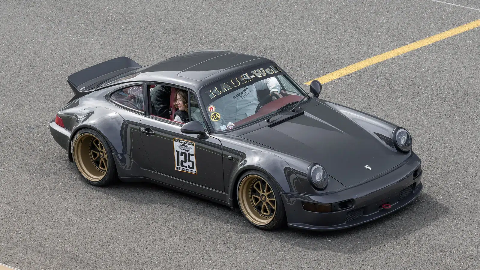 Grey RWB Porsche 964 with gold wheels and a RAUH-Welt windscreen banner on a racetrack pit lane