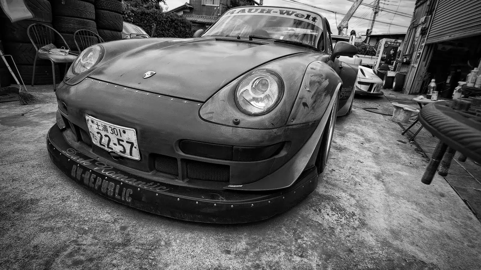 Black and white front view of an RWB Porsche 993 mid-build with a deep front splitter outside a workshop