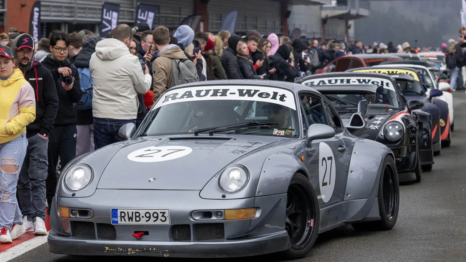 Grey RWB Porsche 993 race car with RAUH-Welt windscreen banner leading a line of RWB cars through a crowd