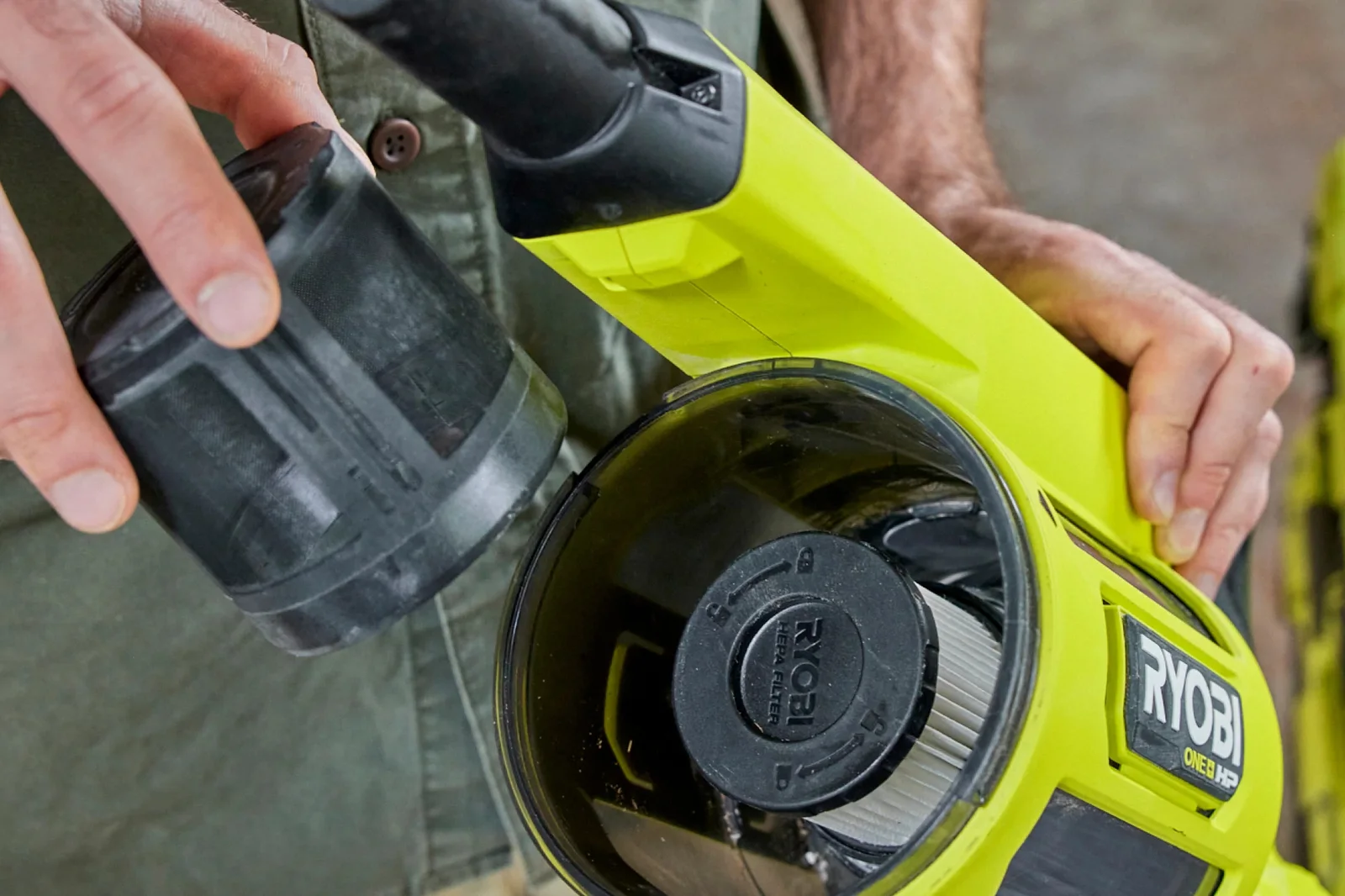 Hands removing the clear 1000mL dust tank from a Ryobi 18V hand vacuum to show the pleated filter