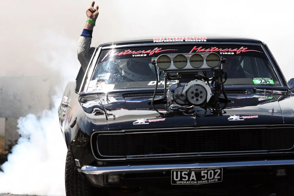 Supercharged black first-generation Camaro laying down a smoky burnout at Summernats in Canberra, driver waving out the window