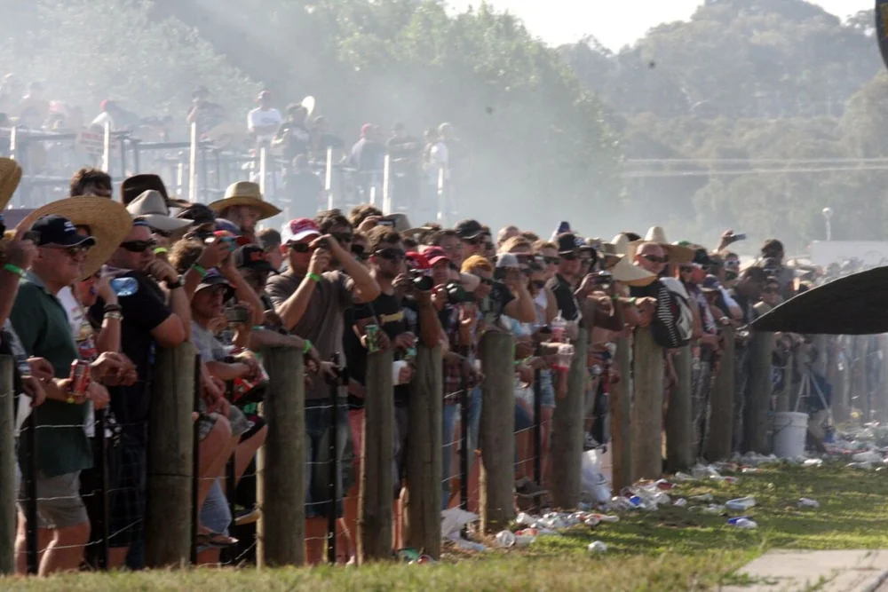 Packed crowd in hats lining the fence of the Summernats burnout pad with tyre smoke drifting over them